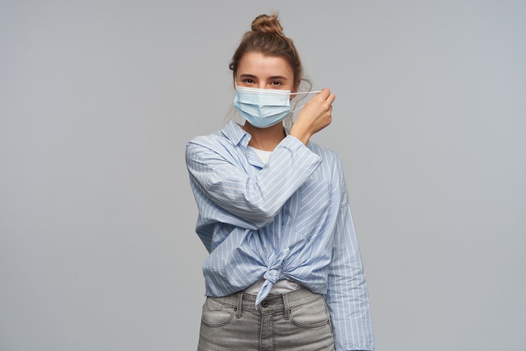 Teenage Girl, Happy Looking Woman With Blond Hair Gathered In Bun. Wearing Striped Knotted Shirt And Protective Face Mask. Takes Mask Off. Watching At The Camera, Isolated Over Grey Background